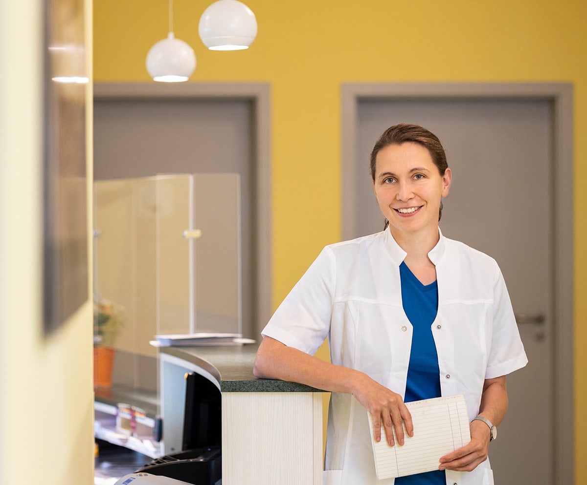 Frau in weißem Kittel mit Patientenkartei mit dem rechten Ellbogen auf der Rezeption lehnend