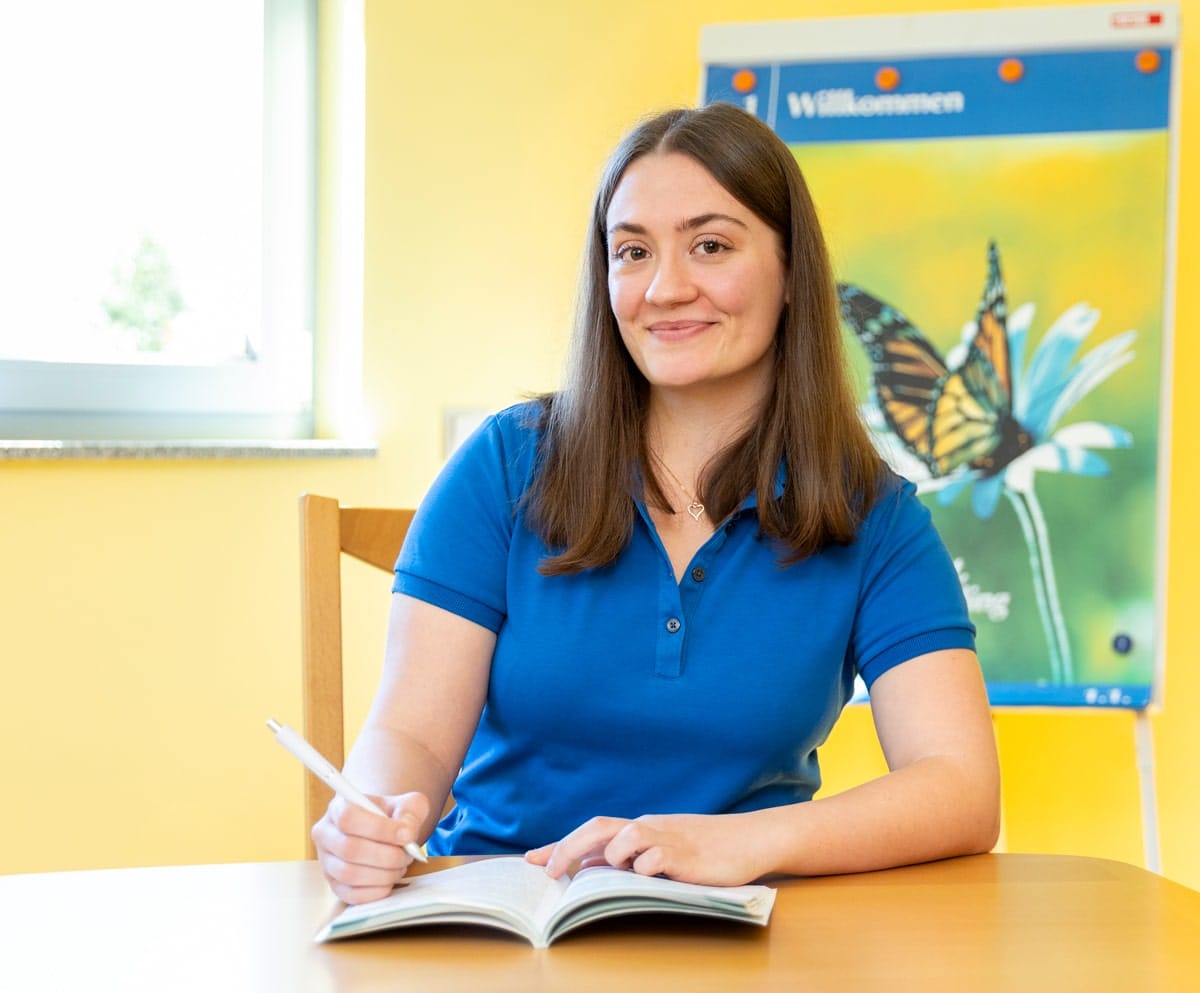 Frau mit blauem Shirt, mit einem Notizheft und Stift sitzend an einem Tisch