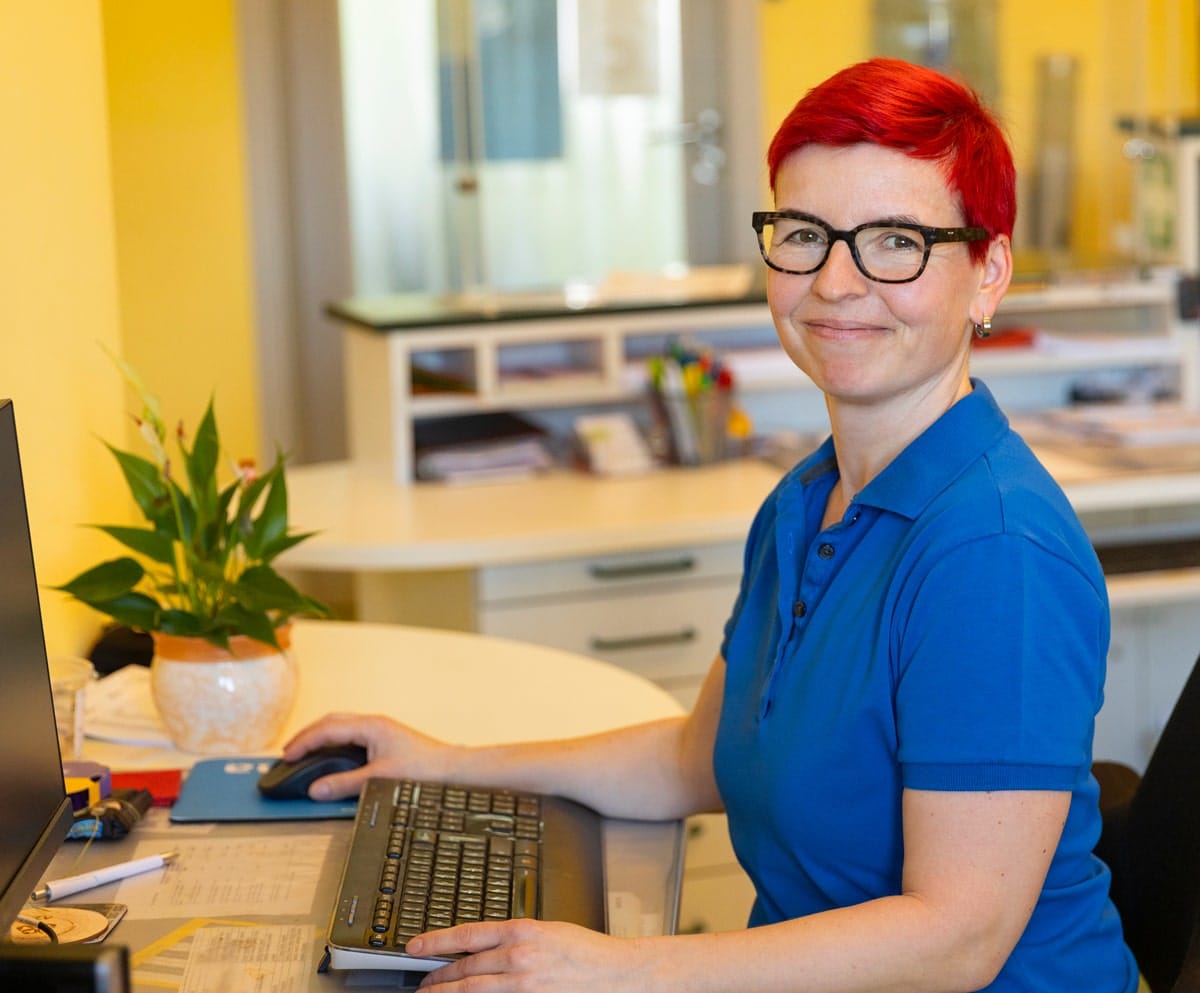 Frau mit blauem Shirt und Brille, sitzend mit einer Computertastatur an einem Schreibtisch