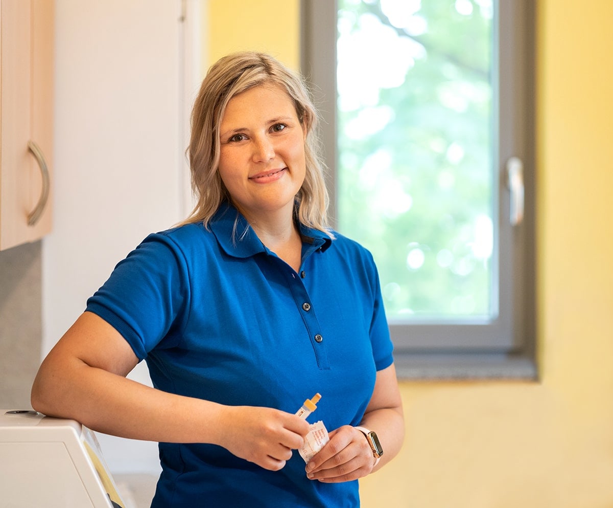 Frau mit blauem Shirt und Kanülen in der Hand an Arbeitsmöbel stehend