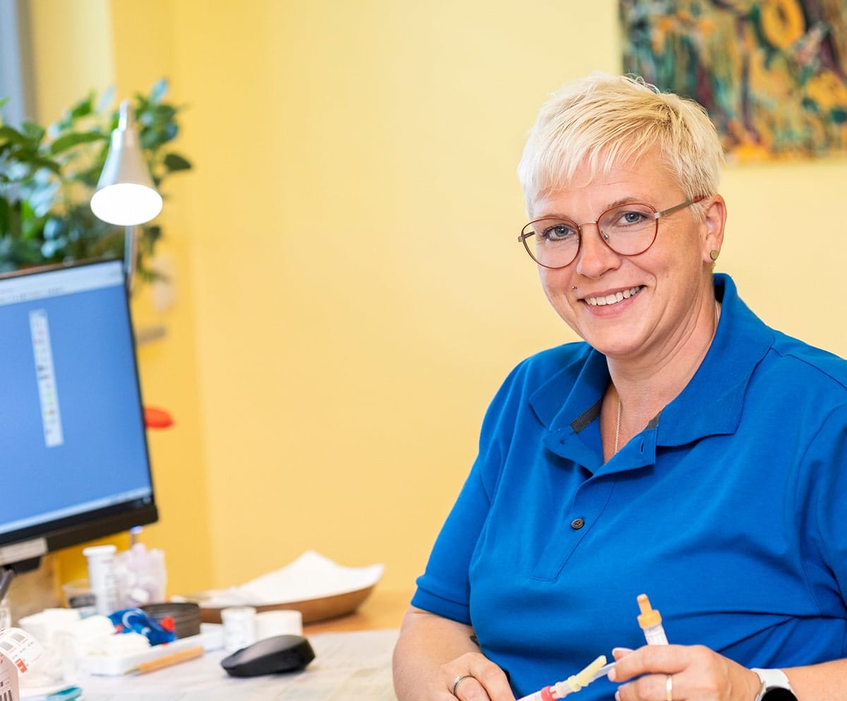 Frau mit blauem Shirt und Kanülen in der Hand, sitzend an einem Schreibtisch